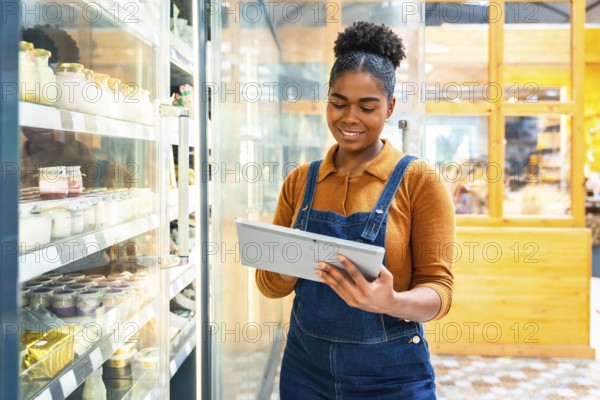 Young supermarket employee using digital tablet checking inventory of dairy products inside of a grocery store refrigerator
