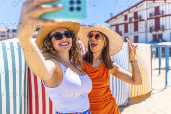 Two cheerful women wearing sun hats and sunglasses, capturing a fun selfie on a sunny beach, radiating joy and friendship