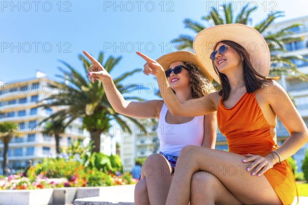 Two happy female tourists wearing sunglasses and summer hats, pointing their fingers at something while enjoying their vacation