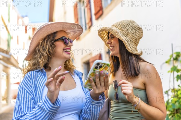 Two happy women wearing summer hats and sunglasses are using a smartphone and laughing while walking down a narrow street on a sunny day