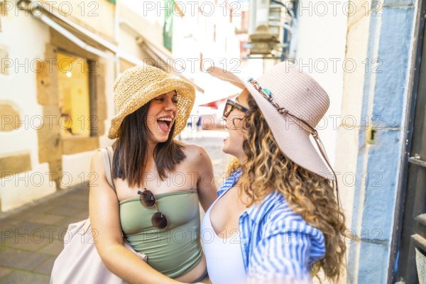 Two happy female tourists wearing summer hats are taking a selfie with a smartphone while walking in a city center during a sunny day