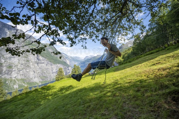 Young man swinging, Blomberg Gård, above the Geirangerfjord, view of idyllic fjord landscape, near Geiranger, Møre og Romsdal, Norway, Europe