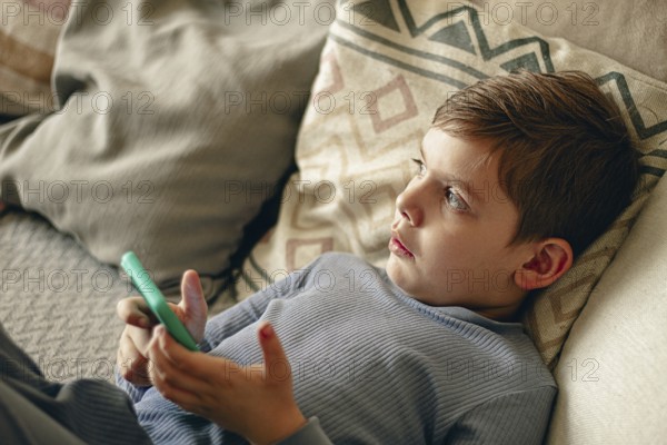 A boy holds a smartphone in his hands, relaxing on the couch, in a cozy living room, natural light, lifestyle