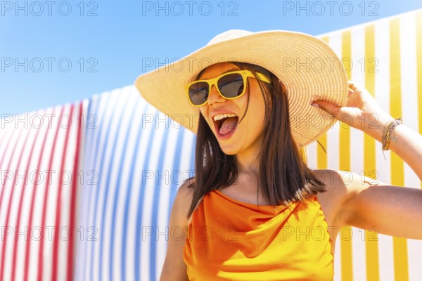 Carefree young woman wearing a straw hat and sunglasses enjoying summer vacation against a colorful striped background