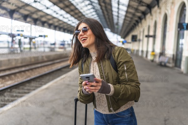 Young woman tourist using her smartphone while waiting for a train at the railway station, smiling and enjoying her travel adventure
