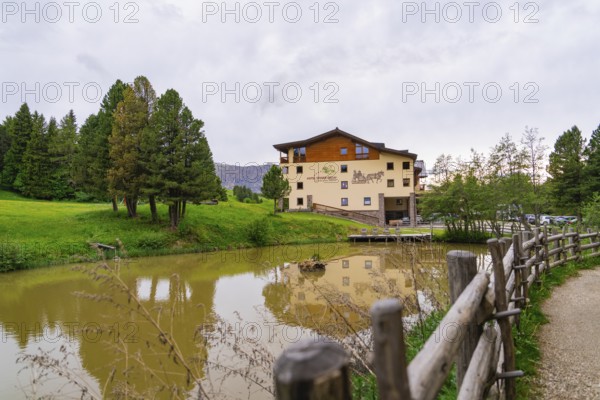 Building at a pond with surrounding trees and a wooden fence under a slightly cloudy sky, Alpe di Siusi, South Tyrol, Dolomites, Italy, Europe