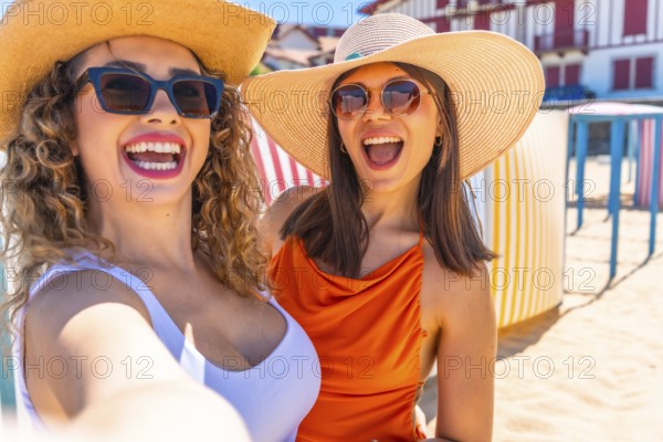 Two cheerful women wearing sunglasses and summer hats are taking a selfie on a sunny beach with colorful beach huts in the background
