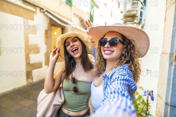 Two joyful women with straw hats and sunglasses are taking a selfie with a cheerful expression, enjoying their summer vacation in a picturesque european town
