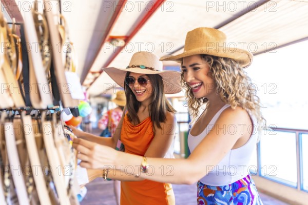 Two smiling women wearing summer hats and sunglasses are choosing souvenirs at a seaside shop while enjoying their vacation