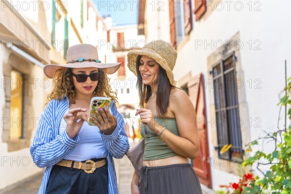 Two female tourists wearing summer hats, using smartphones while walking through charming old town streets, enjoying their adventure together