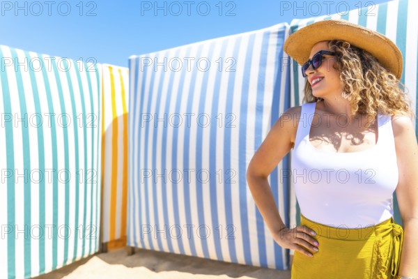 Young woman with curly hair, sunglasses and straw hat posing on beach with colorful striped cabanas in background during sunny summer day
