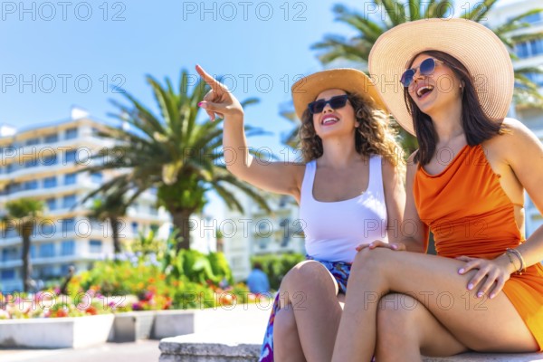 Two happy female tourists wearing summer dresses and straw hats pointing at something while enjoying summer holidays in cannes