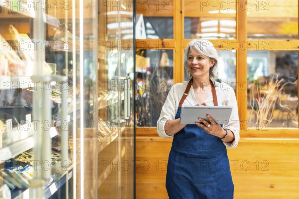 Senior saleswoman wearing apron using digital tablet checking stock availability, working in supermarket
