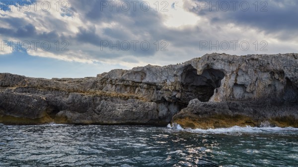 Dramatic rocky coastline with natural arch and cloudy sky over the sea, boat trip around the island of Ortigia, Isola di Ortigia, Syracuse, Sicily, Italy, Europe