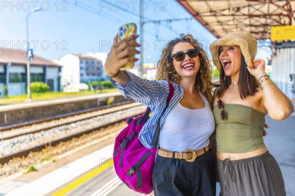 Two happy female tourists are taking a selfie with smartphone at a train station, ready for their summer vacation trip