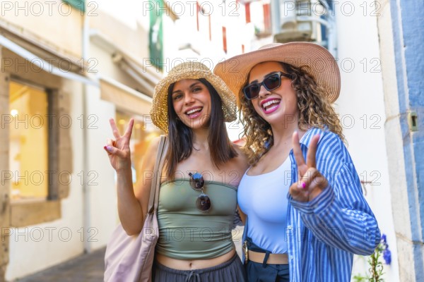 Two happy female tourists wearing summer hats are gesturing the peace sign while enjoying their vacation strolling down a picturesque european street