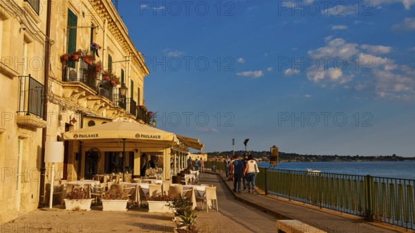 Cosy restaurant on a promenade with sea view. People walking in sunny weather, Ortigia Island, Isola di Ortigia, Historic old town, Syracuse, Sicily, Italy, Europe