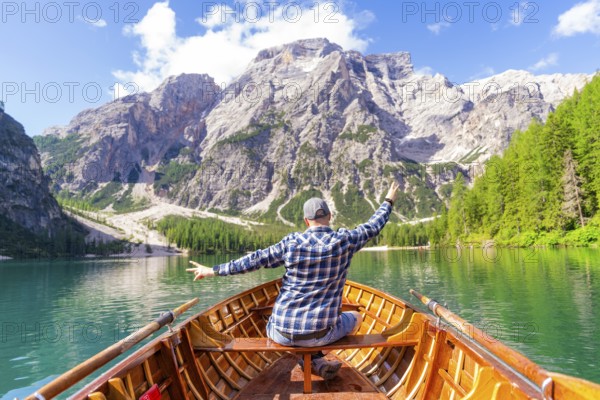 A man sits in a wooden boat and spreads out his arms as he heads towards an impressive mountain, Pragser Wildsee, South Tyrol, Dolomites, Italy, Europe
