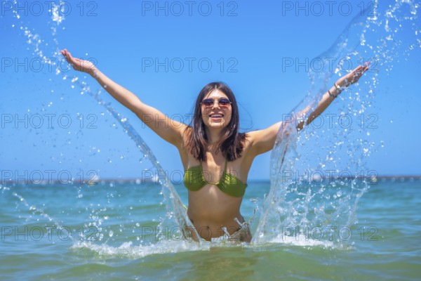 Young woman wearing bikini and sunglasses splashing water with open arms in the sea, enjoying summer vacation on a sunny day