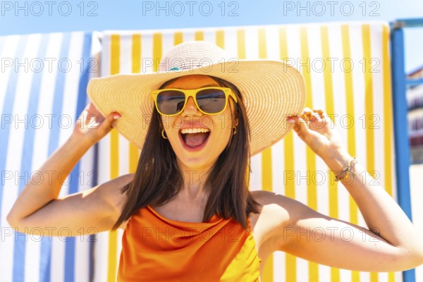 Young woman wearing a straw hat and sunglasses, smiling brightly while enjoying the summer sun, holding her hat with joy and excitement