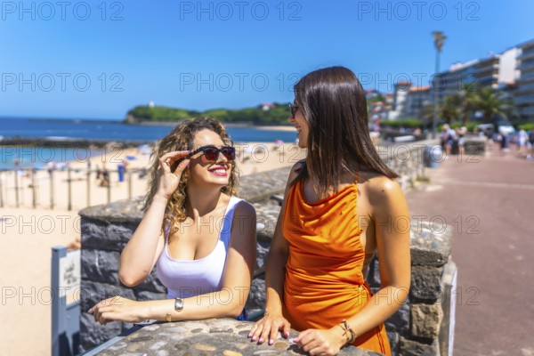 Two cheerful women wearing sunglasses and summer outfits are enjoying their vacation, talking and smiling on a sunny day at saint jean de luz beach