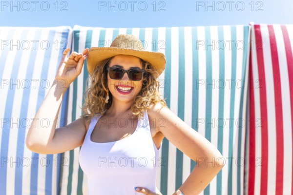 Cheerful young woman with curly hair, wearing sunglasses and a straw hat, smiles while posing confidently against a backdrop of vibrant striped beach towels, enjoying a sunny day at the beach