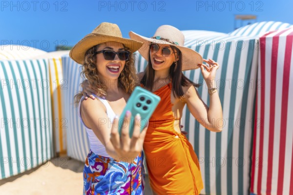 Two happy women wearing summer hats and sunglasses are taking a selfie with a smartphone on a sunny beach, near colorful beach huts
