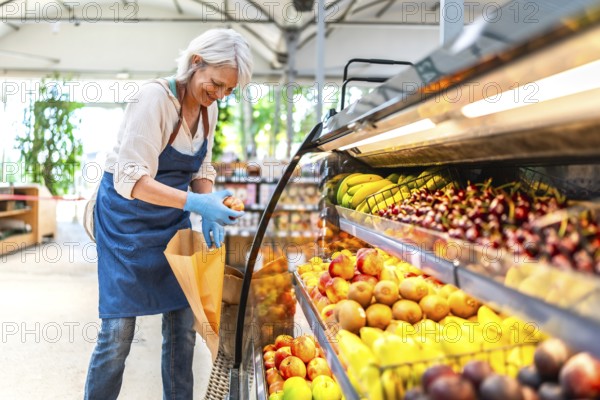 Smiling senior saleswoman wearing gloves, arranging vibrant fresh fruits in a supermarket display case, promoting healthy eating choices