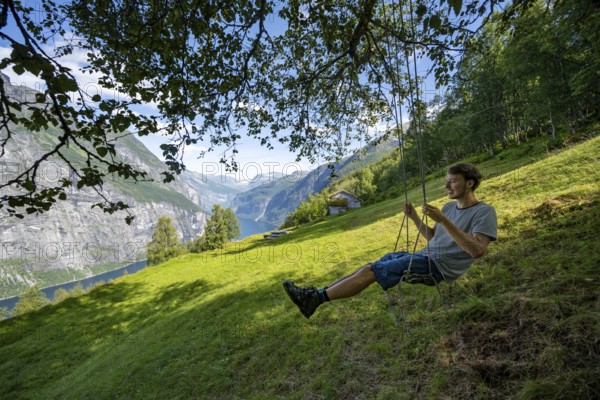 Young man swinging, Blomberg Gård, historic mountain farm with grass roof on a steep mountainside above the Geirangerfjord, view of idyllic fjord landscape, near Geiranger, Møre og Romsdal, Norway, Europe