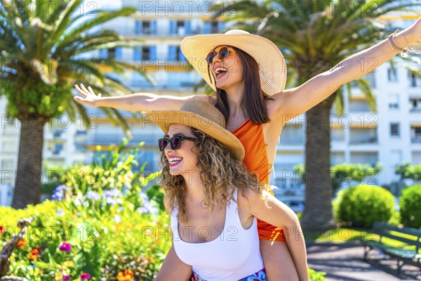 Two cheerful female friends are having fun on vacation in cannes, with one giving the other a piggyback ride, both wearing summer hats and sunglasses