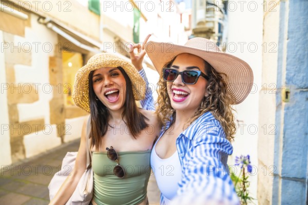 Two cheerful female tourists wearing summer hats and sunglasses are taking a selfie with a cheerful expression while walking down a city street during their vacation