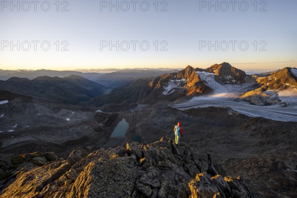 Mountaineer in front of picturesque high mountain landscape at sunrise with alpenglow, blue glacial lakes, glaciers and rocky mountain peaks in the morning light, view at the Becher on mountain peaks Königshofspitz and Sonklarspitze and Schwarzwandspitz with glacier Übeltalferner, Stubai Alps, South Tyrol, Italy, Europe