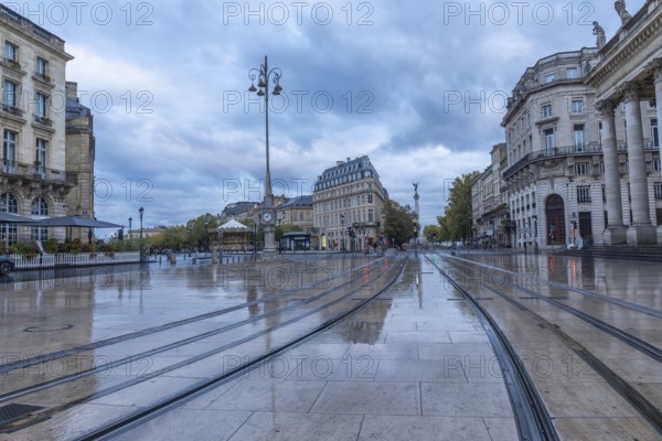Comedie square in bordeaux city center with wet tram tracks reflecting surrounding buildings on a cloudy morning