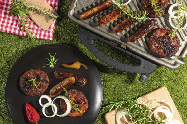 Grilled sausages and burgers on a barbecue and plates with rosemary on a green lawn