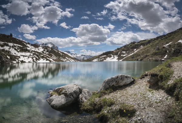 gorgeous mountain lake in the Alps with reflections and snow remnants under a blue sky with white clouds