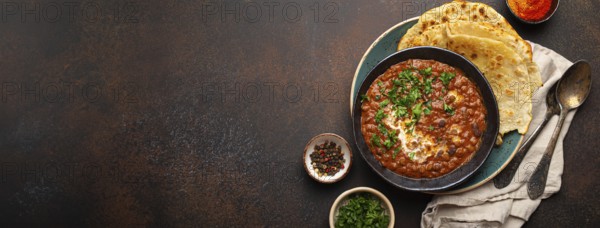 Traditional Indian Punjabi dish Dal makhani with lentils and beans in black bowl served with naan flat bread, fresh cilantro and two spoons on brown concrete rustic table top view. Space for text, food photography, food photography