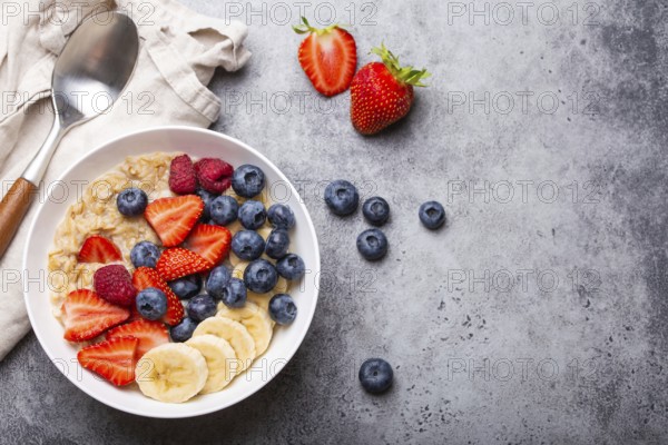 Oatmeal porridge with fruit and berries in bowl with spoon on gray stone background table top view, homemade healthy breakfast cereal with strawberry, banana, blueberry, raspberry. Space for text, food photography