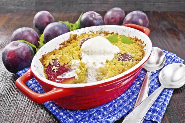 Crumble with plum and ice cream in a red brazier with spoons on a napkin, plums and mint on dark wooden board background