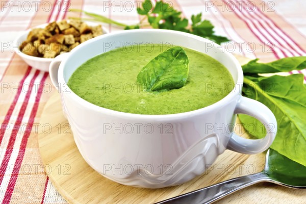 Green soup puree in a bowl with green leaf spinach, spoon on the board, croutons, parsley on a cloth background