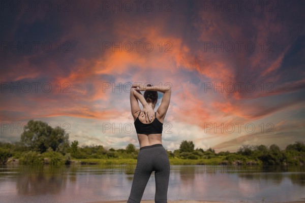Woman performing a yoga stretch by a river at sunset, with a dramatic sky above