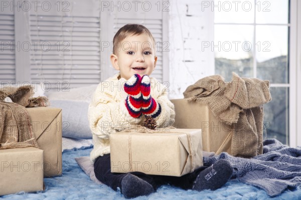 Little funny playful boy a child sits on a bed on Christmas day with gift boxes in white wool knitted jumper and big bright mittens on it and laughs out loud. In interior there is a festive decor