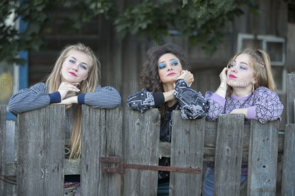Three funny village girls are standing near the old village fence. Bored country girls