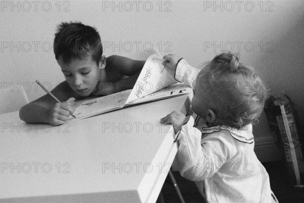A boy sitting at the table and doing his homework. His curious baby sister is standing nearby and holding a page of his book
