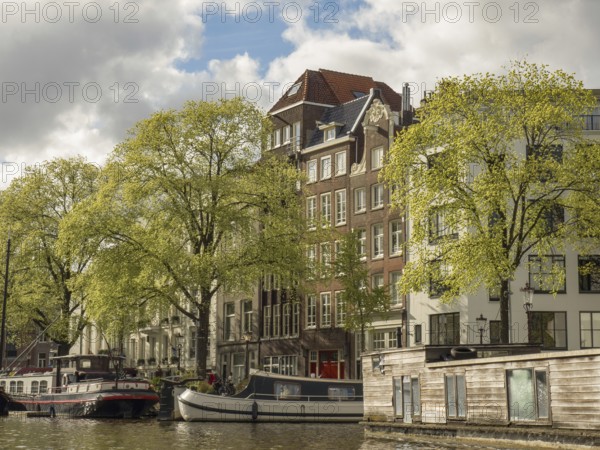 Building fronts along a canal with boats in the water, surrounded by green trees and a cloudy sky, Amsterdam, Netherlands