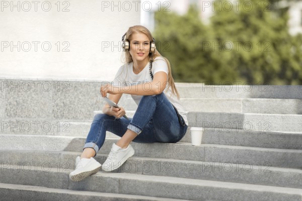 Happy young woman with vintage headphones and a cup of coffee to go, surfing the internet on a tablet PC, listening to music and sitting on a staircase in front of an urban background