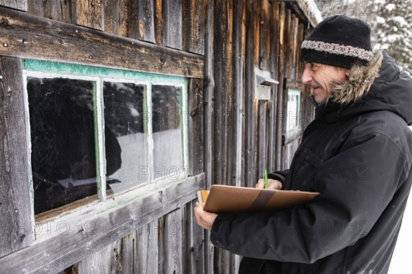 Middle aged home Inspector taking notes on a notebook with leather cover during an outdoor inspection. examining a window of old wooden house