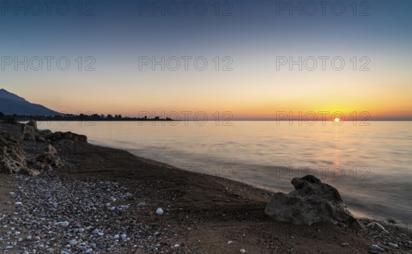 A peaceful sunset over the ocean with silhouette of mountains and pebble beach iwth rock boulders in the foreground