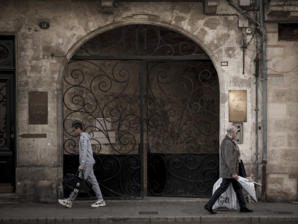 Individuals walk past an arched entranceway with ornate iron gates in central Bordeaux, France, Europe