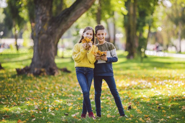 Happy Caucasian siblings enjoying in autumn day in park, sunny weather. Kids having fun holding fallen yellow leaves in nature. Funny twins in autumn park. Autumnal mood. Teenagers posing embracing