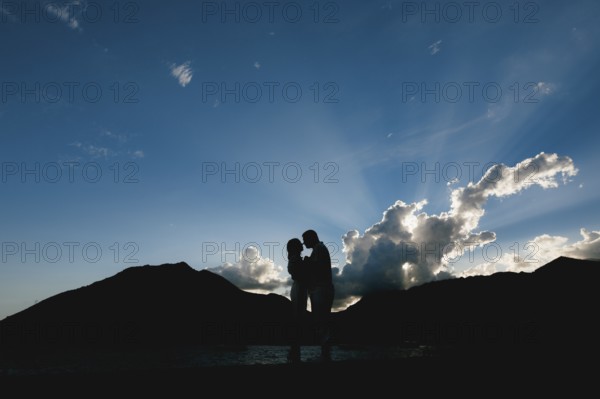 Silhouettes of a man and a woman who hug and kiss at sunset on the seashore against the background of mountains .